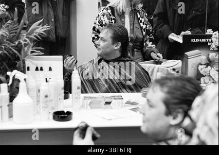 Rob Zorn at hairdresser, Whizgle News, Dutch Desk, The Netherlands, 1950 - 2000 on 15-02-1993. These are the topics in the image. The scene captures a bustling salon environment filled with a mix of activity and anticipation. In the foreground, a man sits in a salon chair, draped in a striped cape that contrasts against his fair skin and dark hair. He appears contemplative, holding a small object in his hand, possibly a product or a tool, as he gazes toward a mirror. The mirror reflects the man's expression, revealing a sense of focus as he engages with the transformation happening. Surroundin Stock Photo
