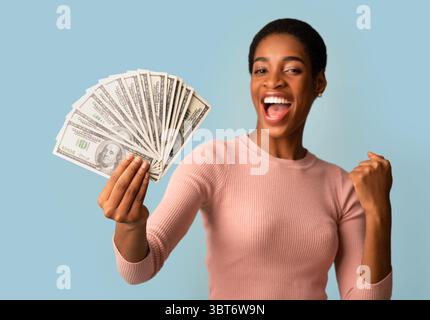 Big Profit. Euphoric Young Black Woman Holding Dollar Cash And Raising Fist Stock Photo