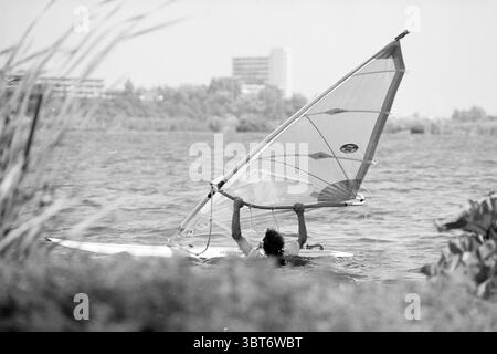 Molenplas Schalkwijk Haarlem The Netherlands, Whizgle News, Dutch Desk, The Netherlands, 1950 - 2000 on 18-07-1992. The image contains these topics. The scene captures a dynamic moment on water, featuring a windsurfer skillfully navigating the waves. The windsurfer is clad in a dark swimsuit, their arms raised high as they grasp the sail, which is predominantly white with some transparent sections, catching the wind. The water beneath them has ripples reflecting the sunlight, creating a shimmering effect that contrasts with the darker tones of the rider. In the background, a blurred skyline re Stock Photo