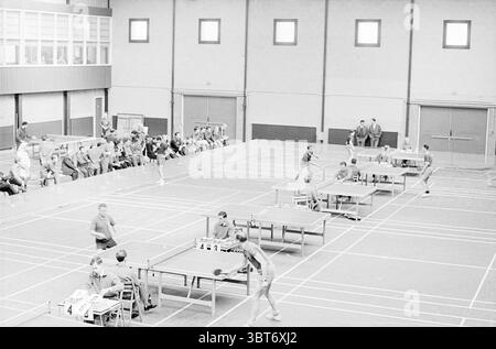 Table tennis tournament Netherlands. Railways Table Tennis, Whizgle News, Dutch Desk, The Netherlands, 1950 - 2000 on 21-04-1964. The image includes these topics. In a spacious indoor venue, multiple table tennis matches are taking place against a backdrop of high ceilings and large windows that allow ambient light to filter in. The overall atmosphere is dynamic and energetic, filled with the sounds of paddles striking balls and the occasional cheer from spectators. The setting features several tables, each surrounded by players in focused stances, demonstrating concentration and athletic prow Stock Photo
