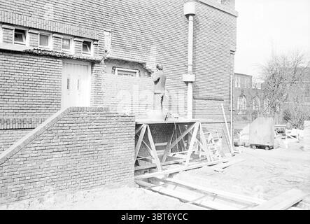 Extra Emmaschool Beverwijk Exterior, Whizgle News, Dutch Desk, The Netherlands, 1950 - 2000 on 17-03-1971. The image shows these topics. A construction site is prominently displayed, featuring a brick wall that showcases a rough patch where restoration work is in progress. At the center, a man stands on elevated scaffolding, meticulously applying mortar to a section of the wall. He is dressed in work attire, indicative of a laborer, focused on his task. The surrounding bricks are a warm reddish-brown, contrasting slightly with the gray cement being used. The scene has a muted quality, as if ca Stock Photo