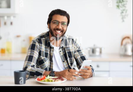 Cheerful middle-eastern man enjoying breakfast, holding smartphone Stock Photo