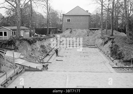 Construction of Denneweg school in Bloemendaal Work in progress Bloemendaal Dennenweg, Whizgle News, Dutch Desk, The Netherlands, 1950 - 2000 on 13-02-1975. These topics are shown in the image. The scene captures an active construction site set against a backdrop of trees, giving a sense of a natural environment juxtaposed with human endeavor. In the foreground, a large, rectangular area, freshly cleared and leveled, appears ready for the foundational work of a building. The ground is covered in light-colored sand, with a few scattered tools and equipment indicating recent activity. Two worker Stock Photo