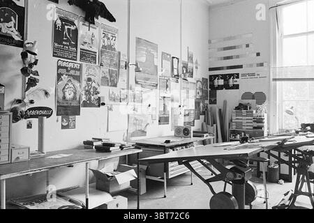 Chris de Jong + Perspect studios Persons, Whizgle News, Dutch Desk, The Netherlands, 1950 - 2000 on 21-04-1977. These are the topics in the image. The scene depicts a spacious, well-lit room that serves as a creative workspace. The walls are adorned with a collage of various posters and artworks, showcasing a mix of styles, colors, and designs. Some posters feature bold, vibrant colors, while others are more subdued, contributing to an eclectic aesthetic. In the foreground, a long, wooden table stretches across the space, serving as a central workspace. Various tools and materials are scattere Stock Photo