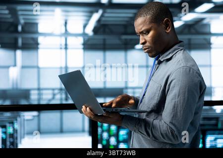 Engineer in data center operating laptop, deploying new server architectures, configuring network virtualization, verifying settings, updating storage arrays, maintaining stable network performance Stock Photo
