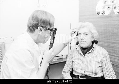 Ophthalmologist Mr. de Jong Doctors, Whizgle News, Dutch Desk, The Netherlands, 1950 - 2000 on 28-02-1978. These are the topics in the image. In a clinical setting, a doctor is examining an elderly patient seated in a chair. The doctor, wearing a white lab coat, is focused intently on the patient, using a handheld eye examination tool to inspect her eye. He has short hair and glasses, which give him a scholarly appearance. The patient has soft, white hair and appears calm, with a slight furrow on her brow that suggests concentration or concern. She is dressed in a striped shirt, the colors mut Stock Photo