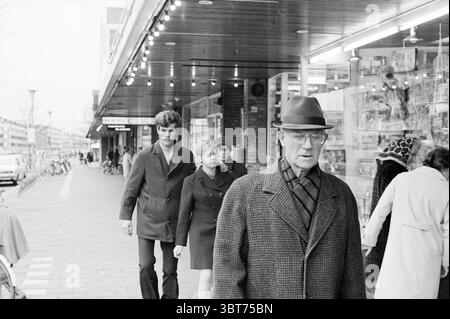 Report about Haarlem Noord Zuid Hollandse Vervoersmaatschappij N.V. NZ Haarlem Grote Markt The Netherlands, Whizgle News, Dutch Desk, The Netherlands, 1950 - 2000 on 16-11-1970. The image includes these topics. In a bustling urban setting, a diverse group of people walks along a sidewalk, flanked by storefronts displaying an array of brightly lit merchandise. The atmosphere is alive with movement and energy. To the foreground, an elderly man wearing a dark overcoat and a wide-brimmed hat strides purposefully, his expression reflecting a mix of contemplation and determination. His glasses catch Stock Photo
