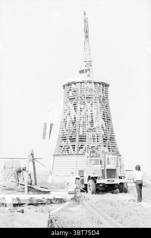 Activities 'De Kat' Uitgeest.' Activities, Whizgle News, Dutch Desk, The Netherlands, 1950 - 2000 on 27-06-1973. These are the elements in the image. The scene depicts a construction site focused on a tall, cylindrical cooling tower, characterized by its lattice-like structure. This tower, with its prominent height, is partially constructed, showcasing both the intricate patterns of the wooden slats and the metallic framework. A crane, positioned nearby, extends toward the top of the tower, where workers are engaged in the assembly process. The crane's arm is prominently displayed, drawing the Stock Photo