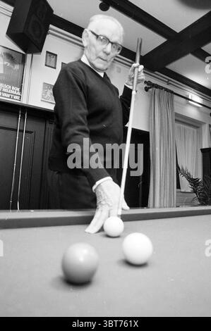 Portrait billiard player., Whizgle News, Dutch Desk, The Netherlands, 1950 - 2000. The image includes these topics. In a lively room with a classic ambiance, an elderly man stands by a pool table, exuding a sense of concentration and focus. He wears a dark sweater and his glasses rest on the bridge of his nose, lending him an air of wisdom. The man is poised with a cue stick, which he grips firmly, preparing to take a shot. His hand is slightly extended towards the table, showcasing a careful movement. The pool table, rich in color, features a smooth surface adorned with three billiard balls: Stock Photo