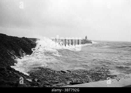 Holes in North Pier IJmuiden Piers and pier construction, Whizgle News, Dutch Desk, The Netherlands, 1950 - 2000 on 30-09-1968. The image contains these topics. The scene captures a dramatic coastal landscape dominated by turbulent waves crashing against a rugged breakwater. The surface of the water is a deep, moody gray, reflecting an overcast sky that suggests a brewing storm. The waves, frothy and white, surge violently as they collide with the concrete structure, sending sprays of water into the air. In the background, a lighthouse stands tall and solitary, its white surface stark against Stock Photo