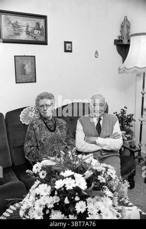 60 years old Willemse family, Whizgle News, Dutch Desk, The Netherlands, 1950 - 2000 on 07-11-1991. These are the elements in the image. In a cozy interior, an elderly couple sits side by side on a dark upholstered sofa, their expressions conveying a sense of quiet companionship. The woman on the left has a thoughtful demeanor, with her gray, tightly curled hair framing her face. She wears a patterned sweater that features a mix of earthy tones—browns, greens, and hints of orange. Beside her, the man has a more neutral expression, dressed in a light-colored vest over a collared shirt. His bald Stock Photo