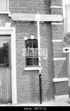 Sign other directions Lisse Borden Lisse, Whizgle News, Dutch Desk, The Netherlands, 1950 - 2000 on 22-06-1984. These topics are shown in the image. The scene features a weathered brick wall, characterized by a rustic red hue, with patches of discoloration indicating age and exposure to the elements. A wooden door is set into the wall, its surface showing signs of wear with a muted brown tone. To the right of the door, a tall, slender pole stands prominently, adorned with directional signs pointing toward different locations. The signs have a monochromatic palette, likely in shades of white an Stock Photo