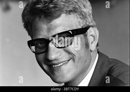 Father J Ruyter Clergy, Whizgle News, Dutch Desk, The Netherlands, 1950 - 2000 on 03-07-1969. These topics are shown in the image. A man with a warm and approachable smile fills the frame, exuding a sense of friendliness. His hair is styled in a light, neat manner, showcasing a greyish hue that contrasts softly with darker roots. He wears large, bold black glasses that dominate his facial features, drawing attention to his expressive eyes. The man is dressed in a suit that appears tailored, with a light or medium-toned fabric that complements his complexion. The backdrop is softly blurred and Stock Photo