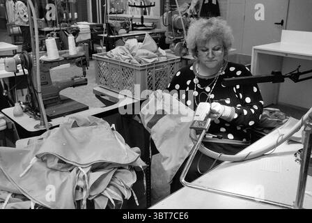 Atelier of clothing company Hensen Haarlem The Netherlands ., Whizgle News, Dutch Desk, The Netherlands, 1950 - 2000. The image contains these topics. In a busy workshop, an individual is seated at a sturdy workstation surrounded by a variety of sewing machines, each displaying spools of thread in vibrant colors. The person is focused on a piece of fabric, handling it with a careful precision that suggests experience. She is dressed in a black garment adorned with large white polka dots, which adds a playful contrast to the muted tones of the workshop. The setting is filled with the textures a Stock Photo