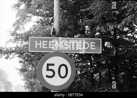 Signs Bloemendaal and Heemstede, Whizgle News, Dutch Desk, The Netherlands, 1950 - 2000 on 25-09-1994. The image contains these topics. The scene presents a tranquil environment, characterized by a blend of natural and urban elements. A metal pole rises vertically, supporting two signs. The upper sign features a rectangular shape with a clean, modern design, its background a bright hue contrasting with bold lettering that stands out prominently against it. Below, a circular sign displays a numerical value, indicating a speed limit, framed by a solid circular boundary. Surrounding the signs, lu Stock Photo