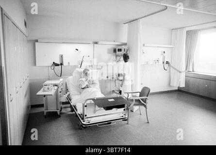 Moving cardiac department Spaarne Hospital Haarlem The Netherlands, Whizgle News, Dutch Desk, The Netherlands, 1950 - 2000 on 08-01-1991. The image includes these topics. The scene depicts a hospital room with a clinical and sterile atmosphere. The walls are primarily painted in soft, neutral tones, contributing to a feeling of calmness. On one side, a patient is seated on a hospital bed, dressed in a light hospital gown. The bed is equipped with clean white linens and various medical devices arranged nearby, indicating that the patient is receiving care. The patient appears contemplative, wit Stock Photo