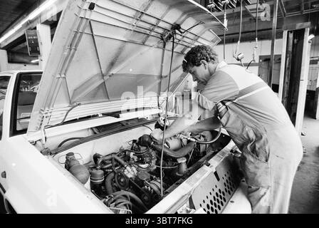 Car mechanic Citroën Haarlem The Netherlands, Whizgle News, Dutch Desk, The Netherlands, 1950 - 2000 on 25-07-1991. These topics are shown in the image. In a busy workshop, a mechanic is engrossed in his task, leaning over the open hood of a vehicle. The scene is defined by a sense of industriousness and focus. The mechanic, dressed in a gray-blue coverall, has a determined expression as he carefully examines the engine components. The vehicle's engine bay is cluttered with various mechanical parts, showcasing an array of metallic colors—silvery grays, deep blacks, and hints of rust. Tools are Stock Photo