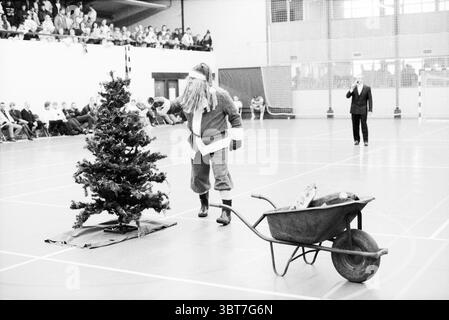 Christmas ball tournament Tybb hall Football, Whizgle News, Dutch Desk, The Netherlands, 1950 - 2000 on 07-01-1989. These are the elements in the image. The scene features a spacious indoor venue, likely a gymnasium or community hall, characterized by high ceilings and large windows that bring in natural light. There is a festive atmosphere permeating the space, underscored by a small evergreen tree, adorned with minimal decorations, standing on a defined area. In the foreground, a person dressed as Santa Claus, complete with a fluffy white beard and red outfit, is engaged in transporting a wh Stock Photo