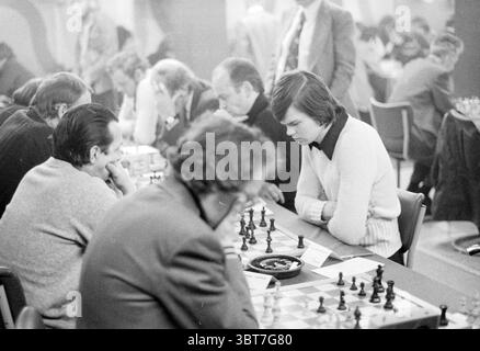 Blast furnace chess tournament Wijk aan Zee, Whizgle News, Dutch Desk, The Netherlands, 1950 - 2000 on 21-01-1976. These are the elements in the image. The scene is a bustling tournament room filled with concentration and tension. At the forefront, several chess boards are set up in a long, rectangular arrangement. Players are seated around the tables, each deeply engrossed in the game before them. A young man in a light-colored sweater leans forward, brows furrowed, analyzing his next move with intense focus. His dark hair is neatly combed, and there’s an air of determination about him. Oppos Stock Photo