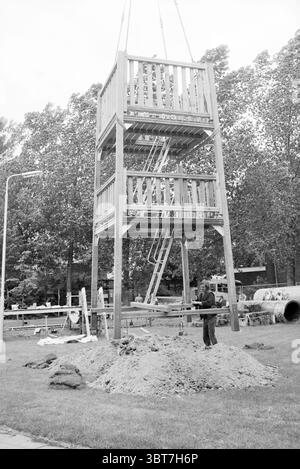 Installing play equipment at the De Ringvaart community center games and toys, Whizgle News, Dutch Desk, The Netherlands, 1950 - 2000 on 05-06-1989. The image shows these topics. The scene depicts a construction site with a large, multi-tiered wooden structure prominently featured at the center. This structure stands tall, elevated above the ground, supported by sturdy vertical beams. The levels are connected by a series of diagonal ladders, suggesting it is designed for climbing or observation. Surrounding the base, a mound of sandy soil gives the impression of ongoing construction work, indi Stock Photo