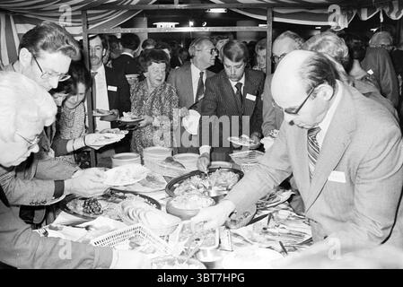 CBR party The Hague C.B.R. Den Haag, Whizgle News, Dutch Desk, The Netherlands, 1950 - 2000 on 20-10-1979. These are the topics in the image. The scene captures a bustling gathering where a diverse group of people is engaged around a large buffet table filled with an array of food items. Various attendees are closely packed together, some leaning over the table, plates in hand, as they serve themselves from a plentiful spread. The buffet features an assortment of dishes, including platters of meats, colorful salads, and stacks of baked goods. The food is arranged attractively, with vibrant col Stock Photo