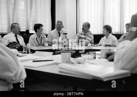 State Inspectorate Meeting, Whizgle News, Dutch Desk, The Netherlands, 1950 - 2000 on 03-05-1990. These topics appear in the image. The scene captures a group of individuals engaged in discussion around a large, rectangular table. The setting appears to be a well-lit conference room, characterized by soft, natural light filtering through large, vertical blinds, creating a warm and focused atmosphere. The people are dressed in business attire, suggesting a formal meeting environment. They exhibit a range of expressions; some are intently listening, while others are animated in conversation, sug Stock Photo