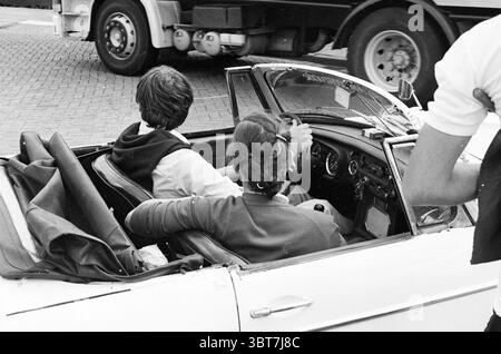 Mosquito Round Mosquito Round, Whizgle News, Dutch Desk, The Netherlands, 1950 - 2000 on 28-07-1982. The image contains these topics. The scene captures a moment inside a classic convertible car, which is a crisp white color demonstrating vintage charm. The interior showcases sleek leather seats, with a polished dashboard displaying elegant gauges and dials, hinting at the car's age and character. In the front seat, a young man and woman are sharing an intimate space. The man, on the left, has dark, tousled hair and is casually dressed in a light-colored shirt with the sleeves rolled up to the Stock Photo