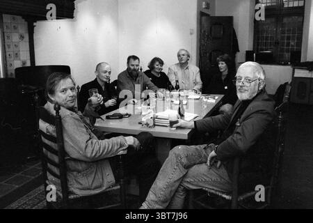 Organization Kunstlijn H'lem Haarlem The Netherlands, Whizgle News, Dutch Desk, The Netherlands, 1950 - 2000 on 31-10-1994. These are the elements in the image. A group of seven individuals is gathered around a large, rectangular table in a warmly lit, intimate setting. The table is cluttered with various objects, including small bottles, drinks, and an array of coasters, suggesting an informal, relaxed atmosphere. The walls of the room are adorned with softly colored paint, and the lighting is warm, creating a cozy and inviting mood. Some sunlight filters through the windows, casting gentle s Stock Photo