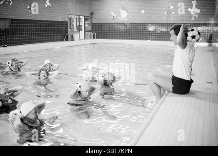 Swimming pool aquagym, Whizgle News, Dutch Desk, The Netherlands, 1950 - 2000 on 05-1989. The image shows these topics. The scene is set in a swimming pool area, characterized by its tiled walls and clear water reflecting overhead lighting. The atmosphere feels energetic and focused, illuminated by bright, artificial lights that create a warm yet vibrant ambiance. In the foreground, a group of several individuals, primarily women, are engaged in an aquatic exercise routine. They wear matching swim caps and appear to be in synchronized motion, lifting objects above their heads with concentratio Stock Photo