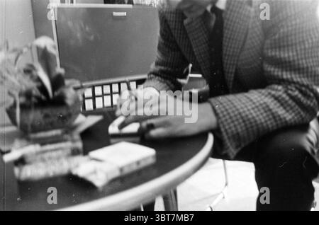 Detail of man at a table., Whizgle News, Dutch Desk, The Netherlands, 1950 - 2000. The image shows these topics. In a softly lit indoor setting, a person is seated close to a round table. The individual is partially obscured, focusing intently on writing. They wear a patterned blazer that adds a touch of sophistication, and their posture suggests concentration and engagement with the task at hand. On the table, a few items are neatly arranged. A small potted plant, vibrant and green, stands out against the more muted tones of the surrounding elements. Next to it, a couple of stacks of what app Stock Photo