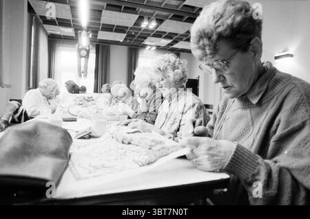Contact for the elderly 't Zijltje, Whizgle News, Dutch Desk, The Netherlands, 1950 - 2000 on 14-10-1991. These are the topics in the image. The scene depicts a group of elderly women engaged in a crafting activity around a long table. The participants, all focused on their work, are seated in a well-lit room. Natural light streams in through large windows, casting a warm glow that contrasts with the cool tones of the walls. Each woman is wearing comfortable clothing, with varying textures and patterns. Some have curly, silver hair, while others have more subdued hairstyles. Their expressions Stock Photo