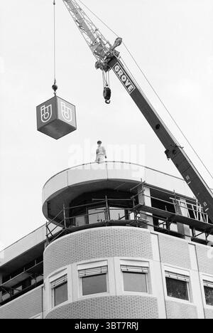 Placing the ABNAMRO logo on the Houtplein building Haarlem Houtplein The Netherlands, Whizgle News, Dutch Desk, The Netherlands, 1950 - 2000 on 29-05-1989. These are the elements in the image. In a scene dominated by a modern architectural structure, a skilled worker is stationed atop a circular, multi-level building that showcases smooth, curved lines. The building's facade is textured with light-colored bricks, providing a contrast against the dark tones of the crane and the man’s work attire. Above him, a towering crane looms, displaying a vibrant yellow and deep red color scheme that draws Stock Photo