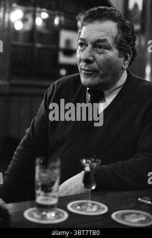 Joachim Noenes pub owner, Whizgle News, Dutch Desk, The Netherlands, 1950 - 2000 on 10-03-1993. The image shows these topics. In a warmly lit, atmospheric interior, a man sits thoughtfully at a wooden table, which has a polished surface reflecting the soft glow of nearby lights. The setting has a classic, cozy bar vibe, with dark wood paneling in the background and blurred circles of light from overhead fixtures creating a bokeh effect. The man, middle-aged with a dignified demeanor, wears a dark sweater over a collared shirt, adding to the relaxed yet polished ambiance. His expression is cont Stock Photo