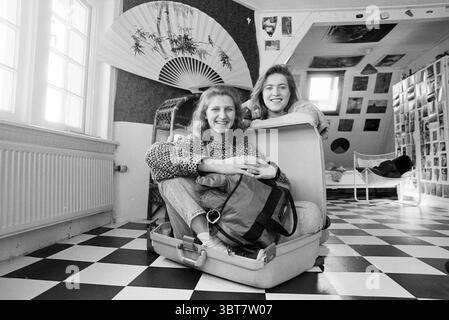 Judith van Beckhoven Saskia van Wabel exhibition in the pegod, Whizgle News, Dutch Desk, The Netherlands, 1950 - 2000 on 09-04-1993. These are the elements in the image. In a spacious room with a unique, eclectic design, two women are seated in an open suitcase, creating a playful and adventurous atmosphere. The room is filled with an array of textures and patterns: the floor is a striking black-and-white checkered pattern that contrasts with the soft light streaming through large windows. The walls feature various decorations, including a large, intricately designed fan that adds a touch of A Stock Photo