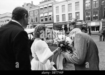Wedding party with Bentley city hall Haarlem Wedding Haarlem The Netherlands, Whizgle News, Dutch Desk, The Netherlands, 1950 - 2000 on 17-04-1986. These are the elements in the image. The scene captures a moment outside in an urban setting characterized by a mixture of old and contemporary architecture. In the foreground, a woman stands gracefully beside a vintage car, her white dress flowing elegantly as she turns to hand a bouquet to an elderly man. The bouquet, colorful with various flowers, contrasts nicely against her attire, adding vibrancy to the composition. The elderly man's posture Stock Photo