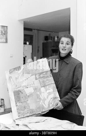 Designer Carolien Crowles Clothing, Whizgle News, Dutch Desk, The Netherlands, 1950 - 2000 on 02-06-1987. The image includes these topics. A woman stands at the center of a well-lit interior space, engaging with her surroundings and holding up a collage of various materials. She has long hair, pulled back, and is dressed in a dark, fitted blazer that contrasts against the lighter tones of the space. Her expression is animated, suggesting enthusiasm or passion for the subject at hand. The collage she presents is a mix of textured elements, possibly made from paper or fabric, with a range of col Stock Photo
