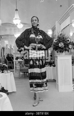 Fashion show Koster, Whizgle News, Dutch Desk, The Netherlands, 1950 - 2000 on 04-03-1996. These are the elements in the image. In the scene, a model stands confidently at the center, showcasing a striking outfit that juxtaposes bold patterns and colors. The garment, a long dress adorned with geometric shapes and contrasting black and white hues, flows gracefully to the floor, complemented by a layered, oversized jacket that drapes elegantly over her shoulders. Her pose is poised, with one hand on her hip, subtly amplifying her stature. A statement necklace draws attention to her neckline, whi Stock Photo