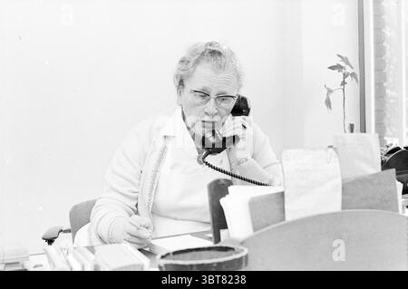 Portrait Sr. Luring Portraits, Whizgle News, Dutch Desk, The Netherlands, 1950 - 2000 on 05-01-1970. The image includes these topics. In a bright, sparsely furnished office, a woman with gray hair styled in soft curls is seated at a wooden desk, which is slightly cluttered. She wears a light-colored cardigan over a collared blouse, and her glasses are perched on her nose as she speaks into a black rotary telephone, holding it with one hand while the other rests on a notepad. The desk is adorned with various items: a stack of papers, a small potted plant with glossy leaves, and a round containe Stock Photo