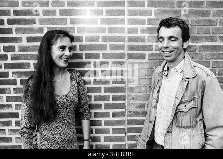 Marleen and Arno organization of the Haarlemmerhout Festival Haarlem The Netherlands, Whizgle News, Dutch Desk, The Netherlands, 1950 - 2000 on 08-06-1997. These are the elements in the image. The scene features two individuals engaging in a lighthearted conversation, set against a textured brick wall that adds depth with its red and brown hues. The wall is composed of neatly arranged bricks, creating a structured backdrop. One person, a woman with long, dark hair cascading over her shoulders, is dressed in a fitted, patterned outfit with a vintage feel, possibly reminiscent of the 70s or 80s. Stock Photo