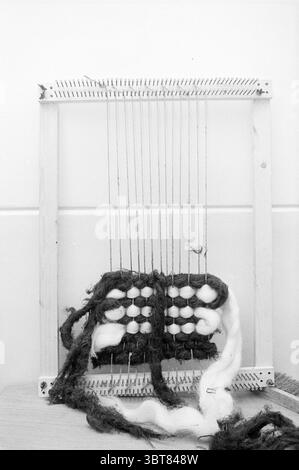 Ms. Jansen weaving Persons, Whizgle News, Dutch Desk, The Netherlands, 1950 - 2000 on 01-10-1973. These are the elements in the image. The scene showcases a simple, handcrafted loom set against a plain, light-colored background, lending a tranquil atmosphere. The loom itself is made of light-colored wood, with vertical threads tautly strung between the top and bottom frames, creating a grid-like structure. Various fibers in natural hues are displayed on the loom, with dark brown and white strands prominently featured, hinting at the textile being created. The contrast between the deep, rich br Stock Photo
