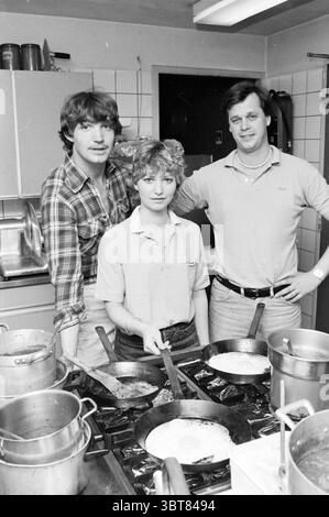 Woman and two men at stove in restaurant kitchen, Whizgle News, Dutch Desk, The Netherlands, 1950 - 2000 on 07-1981. These are the elements in the image. In a bustling kitchen scene, three individuals are the focal point. The two men are situated in the background, while the woman stands prominently in the center, poised and focused on the task. The woman, with short, curly hair, wears a casual, loose-fitting light-colored shirt, giving her an approachable appearance. She holds a spatula, ready to flip the food cooking in front of her. Her expression seems determined, suggesting she is in the Stock Photo