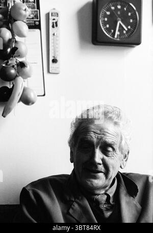 Mr. v.d. Trousers due to robbery Halfweg Halfweg, Whizgle News, Dutch Desk, The Netherlands, 1950 - 2000 on 24-02-1993. These are the topics in the image. In a cozy indoor setting, an elderly man sits comfortably, exuding a warm presence. His gray hair is slightly tousled, framing a face marked by deep lines that tell stories of a long life. A subtle smile graces his lips, hinting at a moment of contentment or nostalgia. The background features a simple yet homey decor. To one side, a cluster of colorful, ripe fruits hangs, adding vibrant hues of yellow, orange, and red against the neutral wal Stock Photo