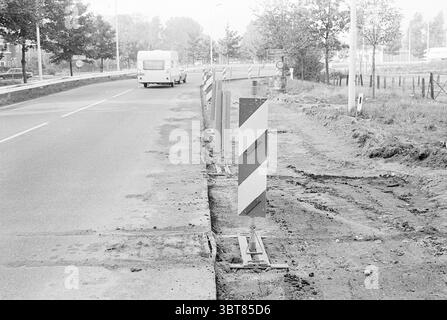 Road works Rijksweg Velsen Roads road construction road planting Work Velsen Rijksweg, Whizgle News, Dutch Desk, The Netherlands, 1950 - 2000 on 27-09-1976. The image includes these topics. The scene depicts a roadway under construction, with a clear focus on the left side where ongoing work is evident. Several concrete barriers are positioned along the edge, marked with diagonal stripes in black and white, indicating caution. These barriers suggest an area that is restricted or altered for safety during the work. The asphalt of the road appears damaged, with sections removed, exposing the und Stock Photo