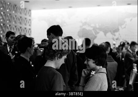 Opening P/A/A. Amsterdam Flowers and flower girls Opening openings inauguration, Whizgle News, Dutch Desk, The Netherlands, 1950 - 2000 on 14-03-1961. These topics are shown in the image. The scene unfolds in a bustling indoor environment, likely a social gathering or reception. The crowd features a diverse mix of individuals, predominantly dressed in mid-20th century attire, reflecting a formal or semi-formal occasion. Men typically wear dark suits, while women showcase a range of styles, including dresses with distinct patterns and textures. In the foreground, two women stand close, engaged Stock Photo