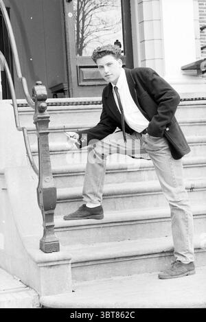 Man on a sidewalk in front of a house., Whizgle News, Dutch Desk, The Netherlands, 1950 - 2000. These topics are shown in the image. The scene captures a young man confidently posed on a set of steps leading to a large entrance. He stands with one foot resting on the lower step, leaning slightly against a decorative railing. His expression is thoughtful, with a hint of rebellion, conveyed through his relaxed posture and slightly tilted head. He is dressed in a classic black blazer over a white shirt, complemented by a black tie. The juxtaposition of the formal blazer with casual denim jeans ad Stock Photo