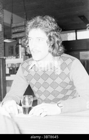 Player George van Haarlem Football Haarlem Football people and trainers, Whizgle News, Dutch Desk, The Netherlands, 1950 - 2000 on 30-01-1975. These topics appear in the image. The scene depicts a young man seated at a bar or café table, exuding a relaxed yet attentive aura. He has curly, dark hair, styled in a way that suggests the era is the late 20th century. A light mustache sits above his upper lip, complementing his youthful features. He wears a patterned sweater that consists of alternating diamonds in muted shades of gray and black, paired with a collared shirt that peeks out from unde Stock Photo
