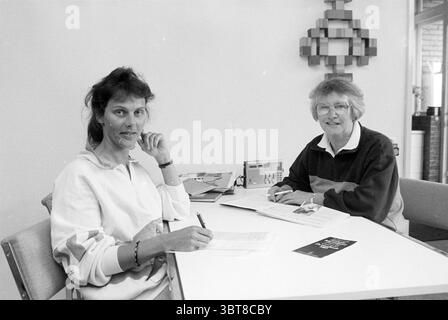 Mrs. Trip from Amnesty International Hillegom Hillegom, Whizgle News, Dutch Desk, The Netherlands, 1950 - 2000 on 22-05-1990. These topics appear in the image. The scene is set in a bright, minimalist interior, characterized by a light-colored wall that enhances the openness of the space. In the foreground, two women are seated at a simple white table. One of them has dark, wavy hair and is wearing a casual white sweatshirt; she holds a pen and appears to be engaged in thought or conversation, resting her chin on her hand. The other woman has short, light-colored hair and is dressed in a darke Stock Photo