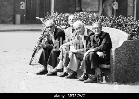 Sun report in Haarlem Report Summer pictures Haarlem The Netherlands, Whizgle News, Dutch Desk, The Netherlands, 1950 - 2000 on 30-08-1979. These topics are shown in the image. In the scene, four elderly men are seated closely together on a stone bench, their postures relaxed yet engaged in conversation. They are dressed in suits, with each man wearing different variations of ties, mostly in subdued colors. Their gray and white hair reflects a wealth of experience, complementing their wise expressions. One of them holds a cane, suggesting a sense of dignity and perhaps mobility challenges. The Stock Photo