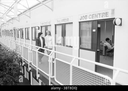 Job center Aalsmeer ., Whizgle News, Dutch Desk, The Netherlands, 1950 - 2000. These topics are shown in the image. The scene features a brightly lit interior space characterized by a modern architectural style. A long, high-ceilinged corridor is visible, lined with multiple offices on either side. The walls display a clean, white finish, contrasting with darker accent colors. Two individuals are walking together along a balcony-like walkway. The first person has curly hair and is dressed in a dark jacket. The second person has long, straight hair and is wearing a light-colored sweater. Their Stock Photo