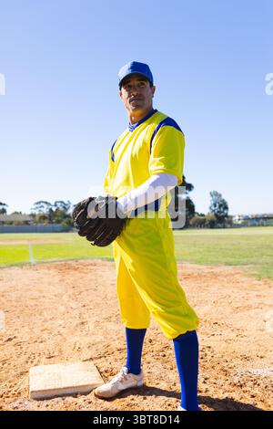 Male athlete standing on infield near base, holding glove, wearing yellow uniform and blue cap Stock Photo